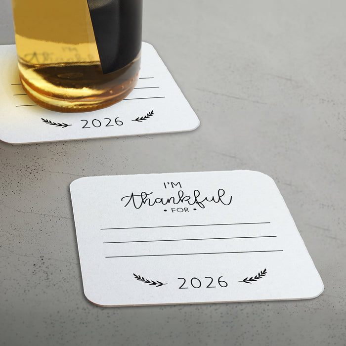 An off-white coaster with a message that says "I'm Thankful for 2026" and a beer bottle on top of it.
