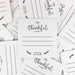 a collection of white coasters with black text and illustrations, arranged in a scattered pattern. The coasters are printed with the text "I'm Thankful for" and lines for people to write on.