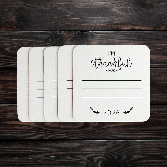 White coasters with black text and designs are shown on a dark wood background. The coasters have the text "I'm Thankful for" and "2026" printed on them.