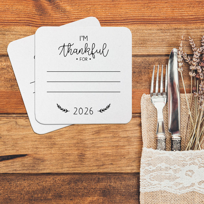 A table setting with coasters that say "I'm Thankful for" and "2026" printed on it, along with a fork and knife placed on a burlap napkin.