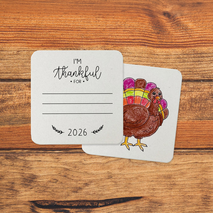 Front and back of coasters shown on an orange, wooden background. Front of coasters show the text Im thankful for, has lines for writing in a response, and current year. Back of coasters feature a turkey illustration for people to color in.