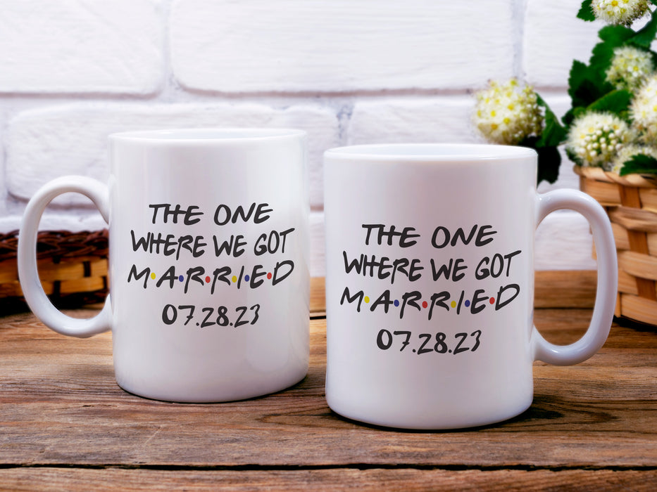 two white coffee tea mugs on wooden table surrounded by cottage core decor, baskets, and plants  mug has tv series Friends Themed design typography that says The One Where We Got Married 07.28.23
