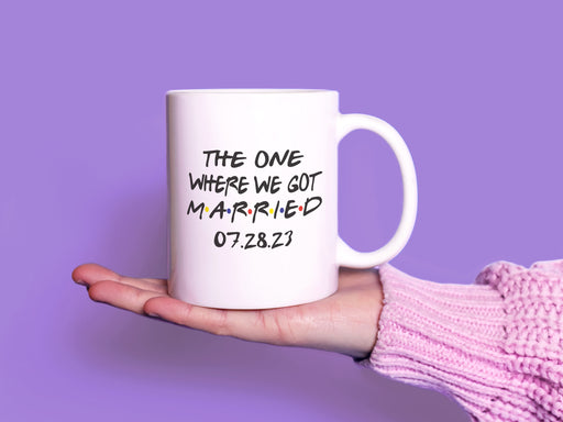 white mug on white table with friends design typography that says The One Where We Got Married 07.28.23 in front of purple background being held by a hand with a pink sweater