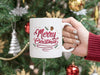 white mug with merry christmas typography artwork being held by a hand with a red sweater in front of a christmas tree decorated with silver, gold, and red ornaments