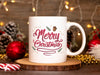 white mug with merry christmas typography artwork on a wooden coaster surrounded by christmas items such as a candy cane, red ornament, pinecones, golden beads, and christmas lights