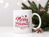 white mug with merry christmas typography artwork on white table next to pine leaves and pine cones with christmas lights In the background