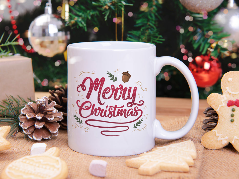 white mug with merry christmas typography artwork surrounded by christmas cookies, pine cones, pine leaves in front of a christmas tree decorated by silver and red ornaments