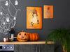 2 frames of wall art with halloween designs on a kitchen wall with a countertop with pumpkins and spider ghost decoration. art designs include; a cat dressed as a ghost carrying a pumpkin, and a skeleton hand
