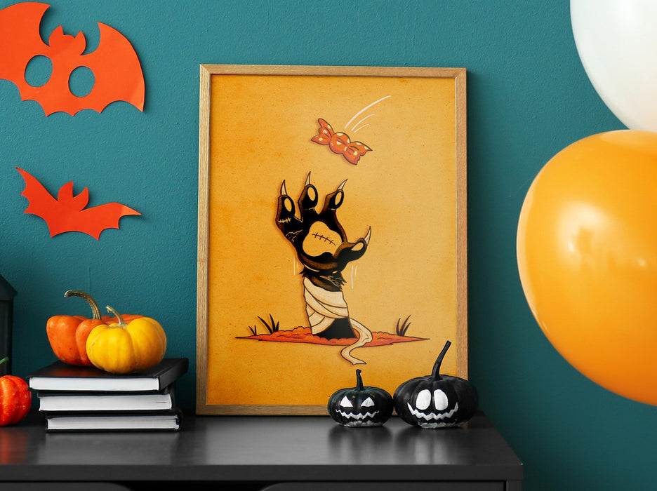 halloween zombie cat paw art print in wooden frame leaning against teal wall sitting on black dresser counter surrounded by halloween decor such as orange and white balloons, bats, pumpkins, jack-o-lanterns, and candles