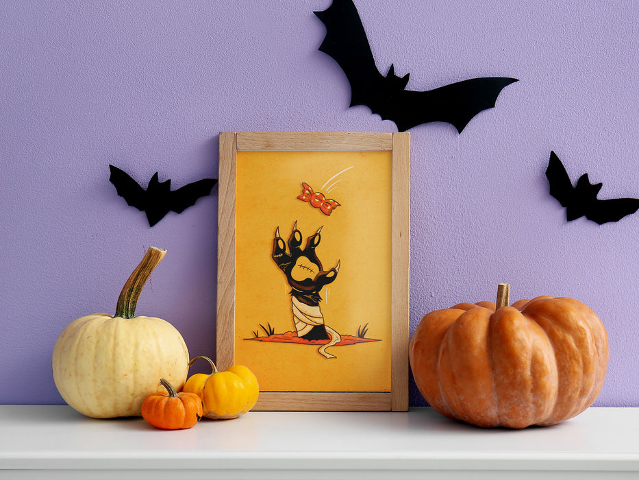 single photo wooden frame of retro halloween artwork of a zombie mummy cat paw grabbing a candy against purple background with pumpkin and bat decoration on top of white countertop