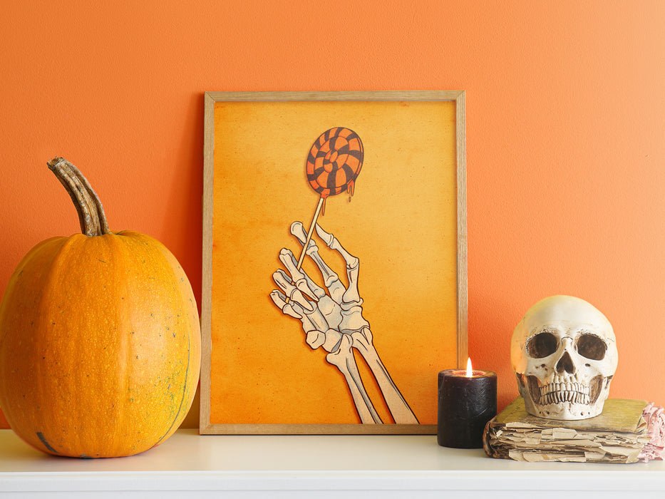 halloween art print of skeleton hand holding a lollipop in a wooden frame in front of an orange wall ontop of a white countertop surrounded by halloween decor such as pumpkins, candles, and a white skull
