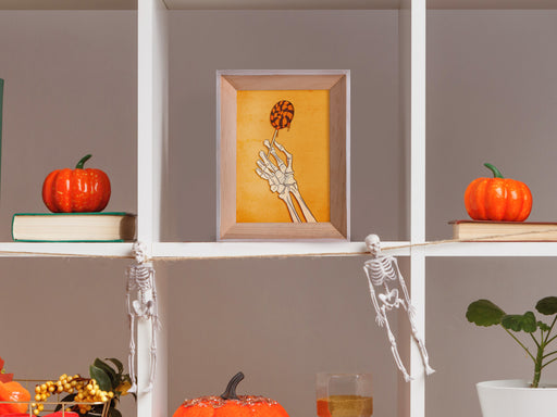 single wooden photo frame with Halloween design of a skeleton hand holding a lollipop with a yellow background with vintage retro patterns on white shelves surrounded by decor such as pumpkins and skeletons
