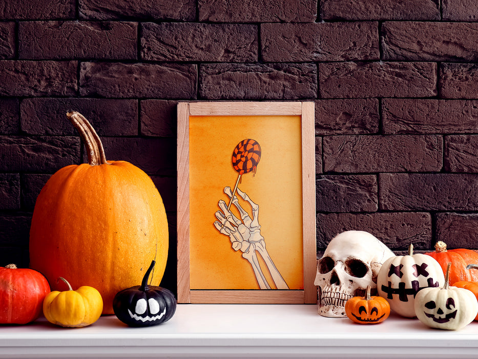 single wooden frame with halloween art of a retro vintage skeleton hand holding a black and orange lollipop against a yellow background against a dark brick wall surrounded by pumpkins, jack o lanterns, and a skull