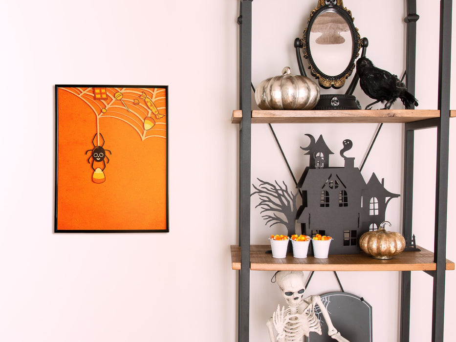 Single framed wall print of spider holding candy corn with web full of candy and orange background against a white wall next to decorated shelves with halloween pumpkins candy corn and a skeleton