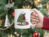 white mug with mouse decorating christmas tree artwork being held by a hand with a red sweater in front of a christmas tree decorated with silver, gold, and red ornaments