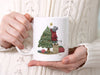 white mug with mouse decorating christmas tree artwork being held by a woman in a white knit winter sweater