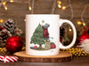 white mug with mouse decorating christmas tree artwork on a wooden coaster surrounded by christmas items such as a candy cane, red ornament, pinecones, golden beads, and christmas lights