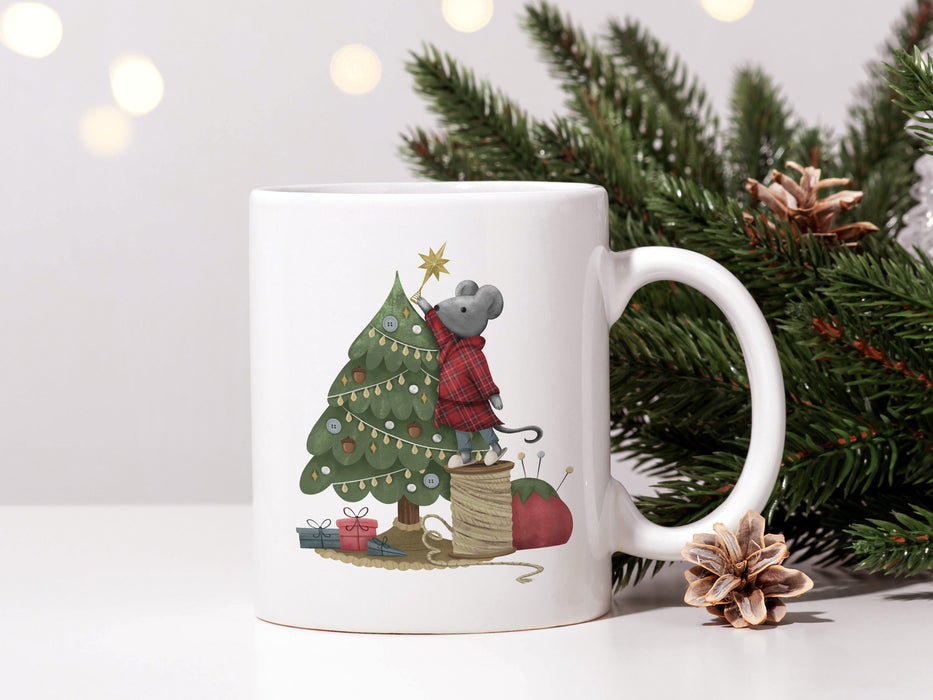 white mug with mouse decorating christmas tree artwork on white table next to pine leaves and pine cones with christmas lights In the background