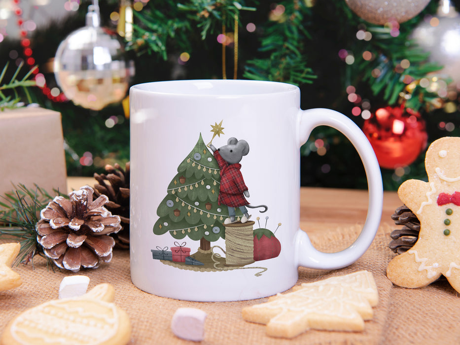 white mug with mouse decorating christmas tree artwork surrounded by christmas cookies, pine cones, pine leaves in front of a christmas tree decorated by silver and red ornaments