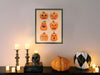halloween art print in frame hung on white wall depicting two rows of pumpkins with different faces, hung over counter surrounded by halloween decor such as pumpkins, candles, and a black skull
