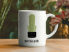 white mug with don't be a prick typography design with cactus graphic on wooden table with flowers in front of it and various house plants in the background