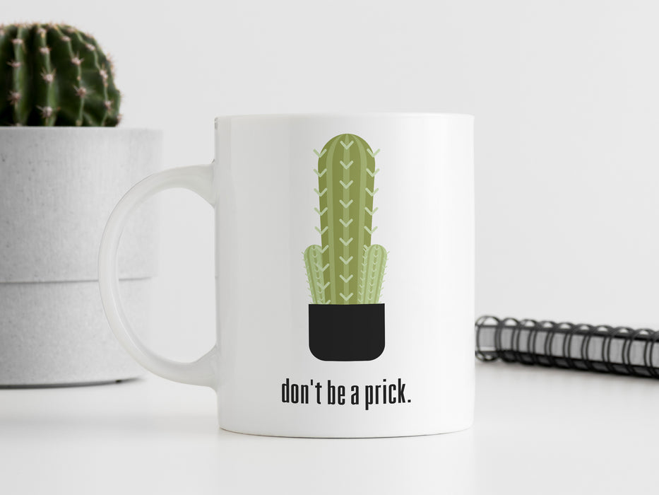 white mug with don't be a prick typography design with cactus graphic on white table in front of notepad and a potted cacti in front of a white wall