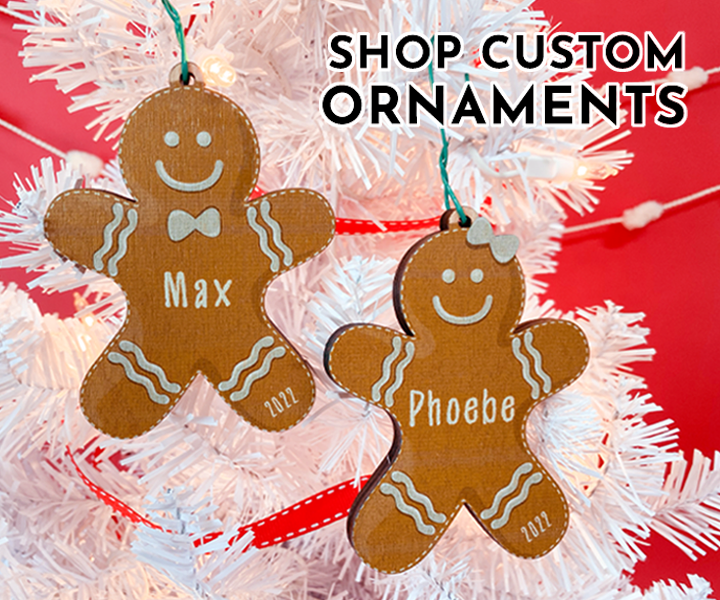 Text: Shop Custom Ornaments Image: Two gingerbread people ornaments are shown hanging on a white Christmas tree. The background behind the tree is red.