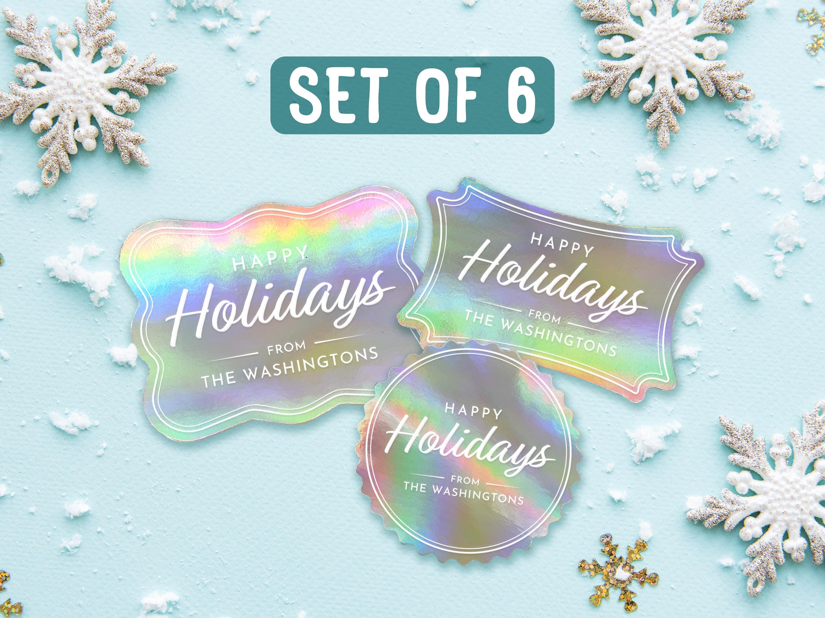 Text reads, Set of 6. Three iridescent foil cardstock Happy Holiday gift tags are shown on a light blue surface surrounded by snowflakes.