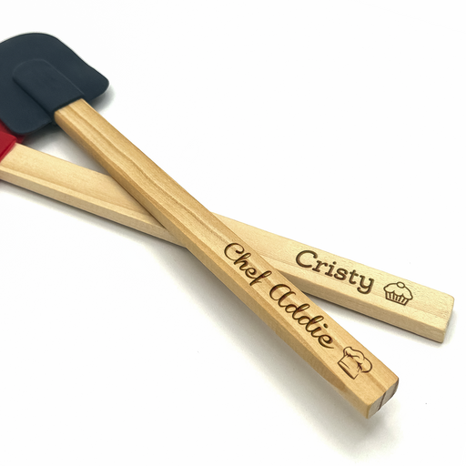 two wooden spatulas in the colors midnight and red on white background with engraving on handles, one says Cristy with cupcake icon and the other says Chef Addie with chefs hat icon