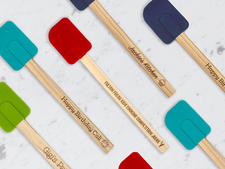 different colored spatulas laid out on a marble table with red, teal, green, and midnight colors with engravings on wood handles that say Jackie's Kitchen  Fulton Fresh Kids Cooking Competition Judge Happy Birthday Cali and Gigi's Patisserie