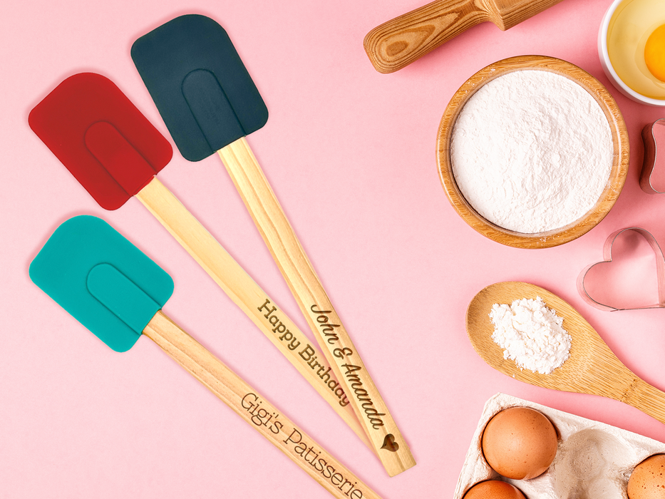 three silicone spatulas with wooden handles in the color red, midnight, and teal with engravings on handles that say John & Amanda With a heart icon, Happy Birthday, and Gigi's Patisserie Spatulas are on pink background surrounded by baking supplies