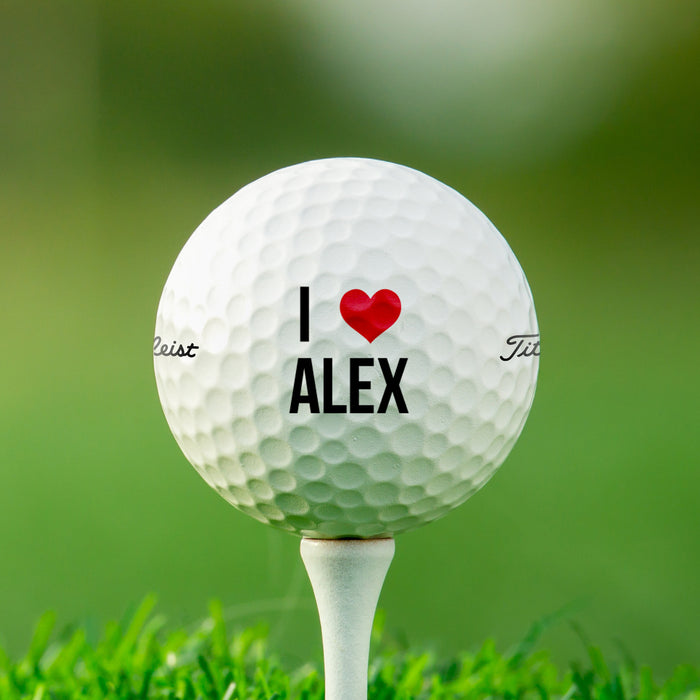 single white titleist golf ball with customizable personalized name and heart design on white golf tee on golf course grass background