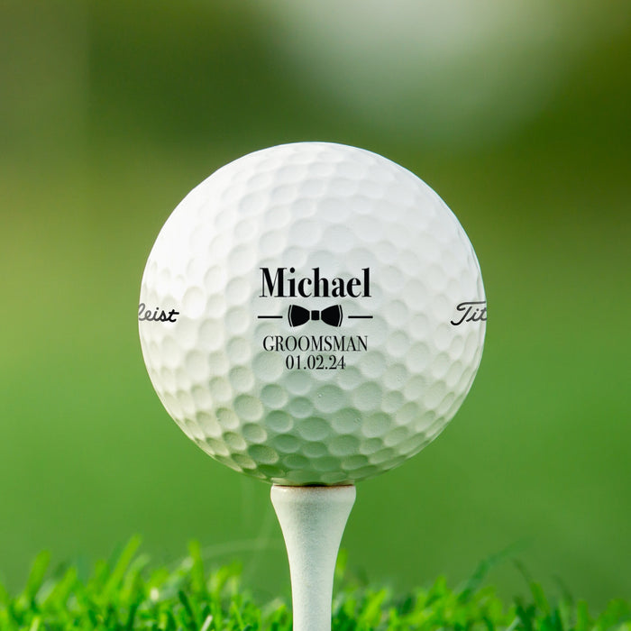 single white titleist golf ball with customizable personalized groomsmen bowtie design on white golf tee on golf course grass background