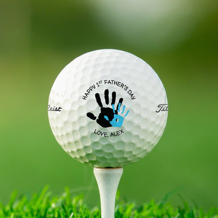 single white titleist golf ball with custom personalized name happy 1st fathers day handprint design on white golf tee with golf course grass in the background