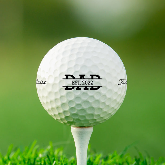 single white titleist golf ball with custom personalized established dad design on white golf tee in front of green golf course grass background 