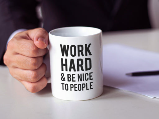 white work hard and be nice to people mug being held by a businessman at a work desk next to a paper and pen