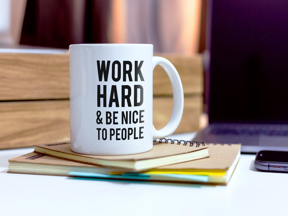 white work hard and be nice to people mug on desk on top of beige notebooks next to laptop and other desk supplies