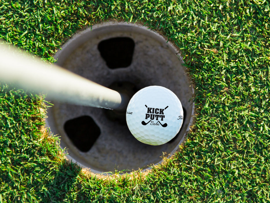single white titleist golf ball with kick putt babe design in golf hole next to pole surrounded by green golf course grass