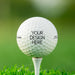 single white titleist golf ball with custom design here text on white golf tee on top of golf course grass