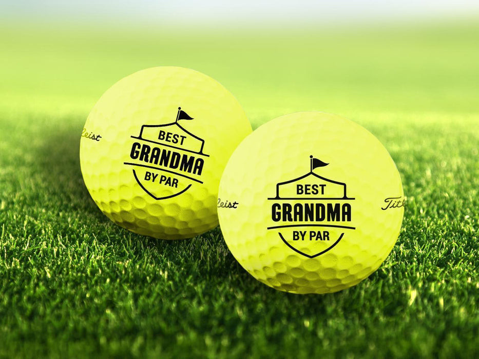 Two personalized yellow golf balls with a Best Grandma By Par design printed on them, placed on a grassy field.