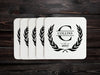A stack of coasters are spread out on a dark wooden table. Coasters say Collins Est. 2017 with laurel design.