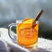 A glass mug filled with a warm beverage, likely tea, and garnished with an orange slice and a cinnamon stick. The mug has a personalized message engraved on it, which reads "Happy Mother's Day SCARLET, 2026".