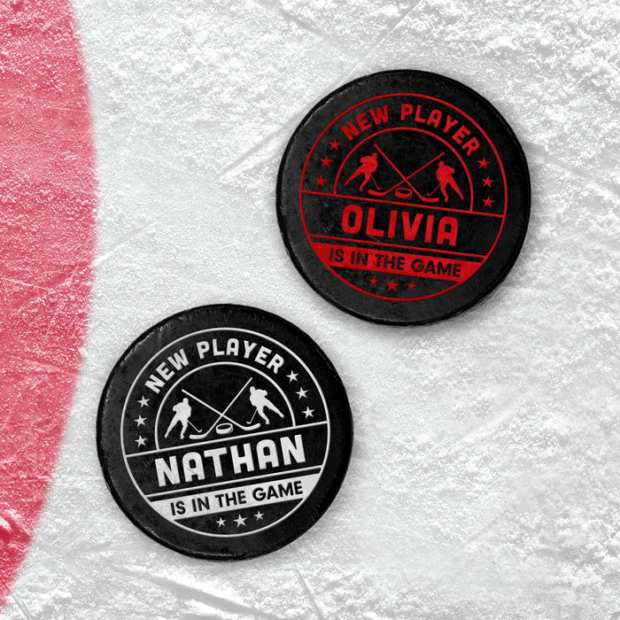 Two personalized hockey pucks with a UV printed “New Player” birth announcement design, shown in red and white on an icy surface.