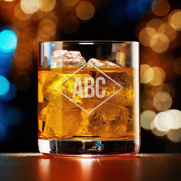 Elegant product photo of the engraved whiskey glass against a warm bokeh background. The diamond monogram is centered and clearly visible.