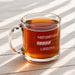 A glass mug filled with a warm beverage, such as tea or coffee, sits on a wooden surface. The mug has a motivational message engraved on it, which reads "Motivation Loading".