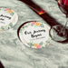 Personalized floral Save the Date coaster displayed beside a wine glass. Features a floral wedding design with custom details.