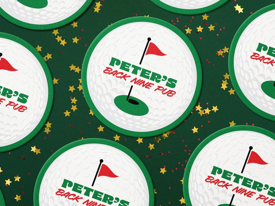 Custom round pulpboard coasters are shown spread out across a dark green surface covered in red and gold star confetti. The custom coasters features a green border, golf ball, a red flag, and golf hole illustration and bold text across the center that can be customized with a name and lounge name.