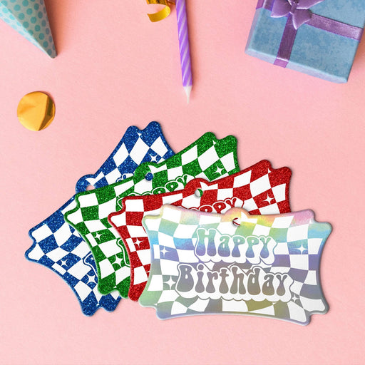 Four groovy birthday tags in metallic rainbow, glitter blue, glitter green, and glitter red lying beside colorful gifts and party decorations.