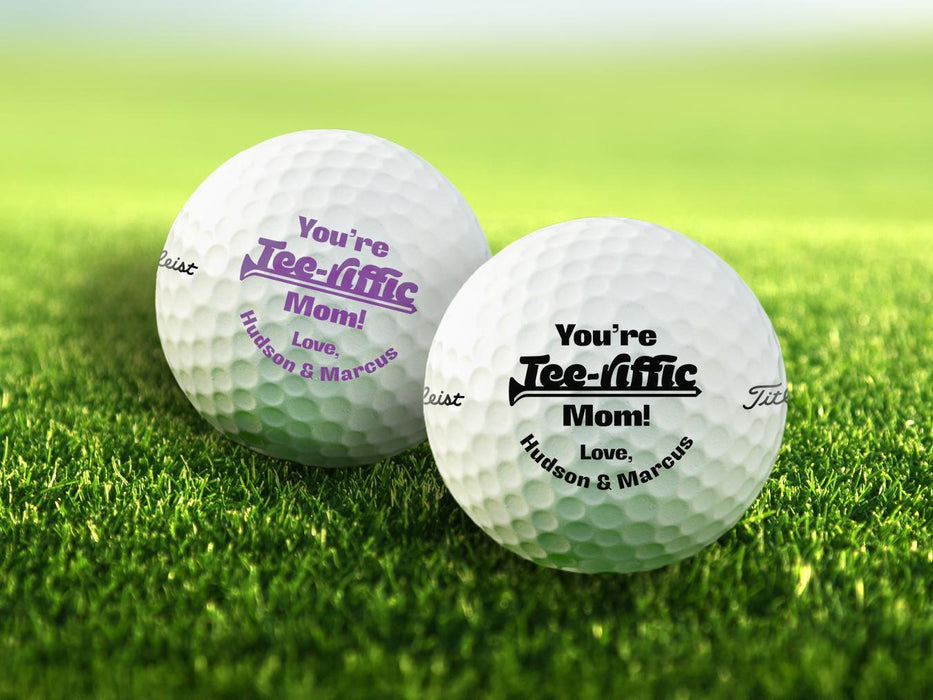 Two personalized golf balls with You're Tee-riffic Mom designs printed on them, placed on a grassy field.