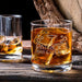 Lifestyle side-angle image of the whiskey glass with whiskey and large ice cubes in a warm home bar setting. The engraved design adds a custom, upscale touch perfect for gifting.